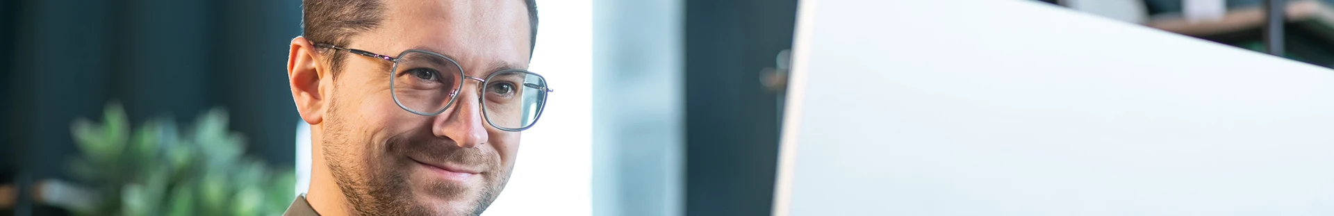 Man with glasses smiling while looking at a computer monitor in a bright office setting.